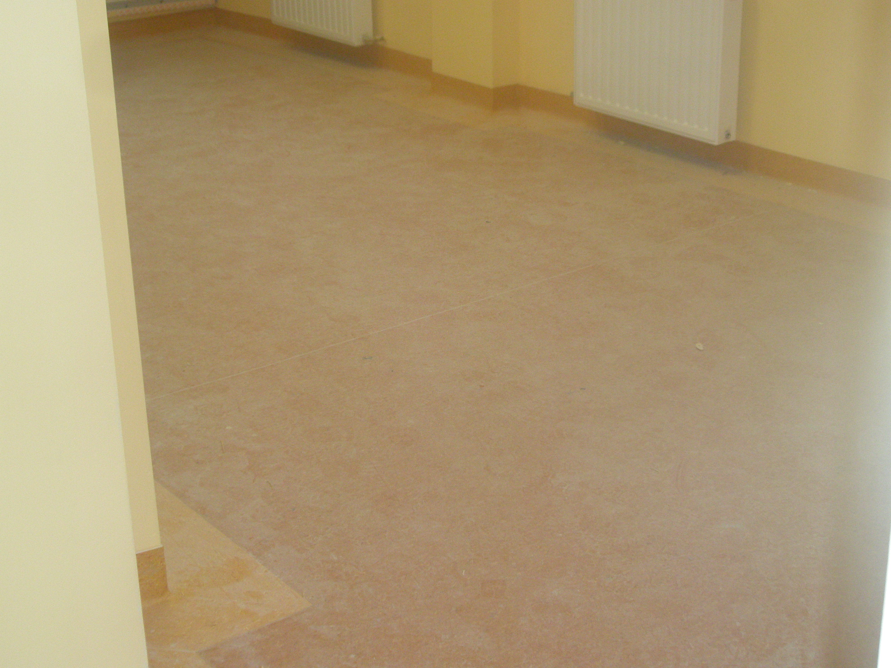 Jasnobrązowa podłoga wykończona listwami przypodłogowymi, żółte ściany z białymi grzejnikami, widok z rogu pokoju.